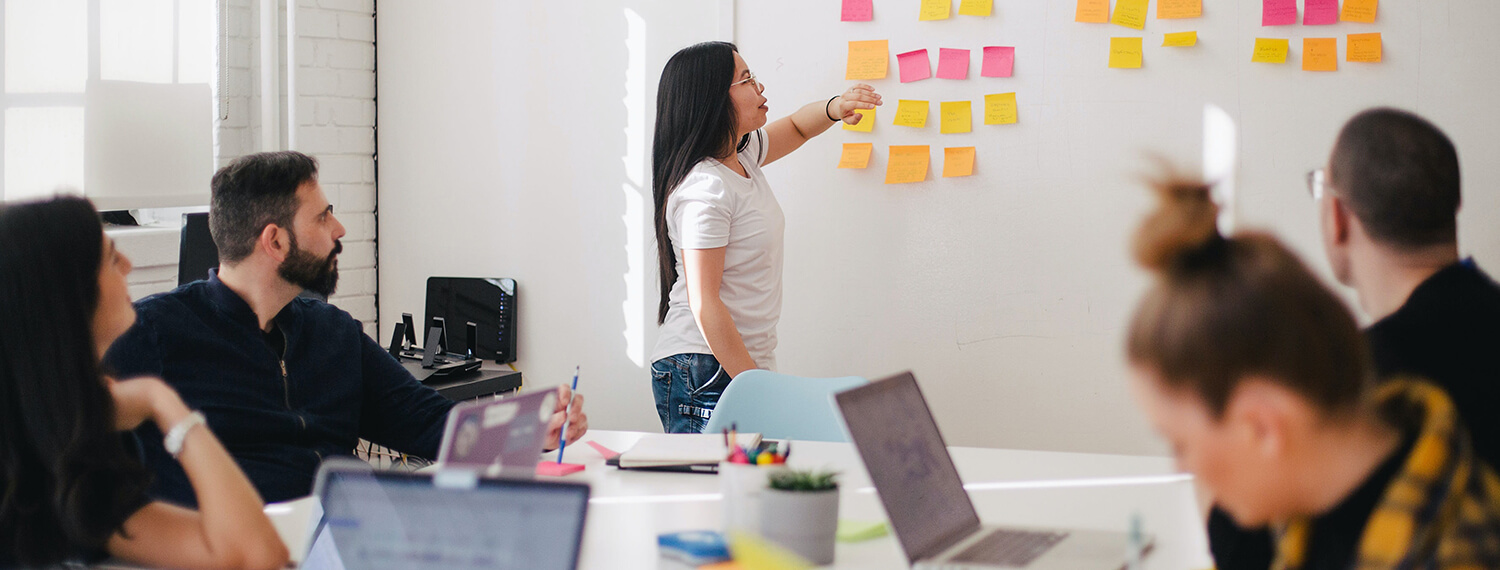 Corporate team meeting with a presenter pointing at a whiteboard with notes